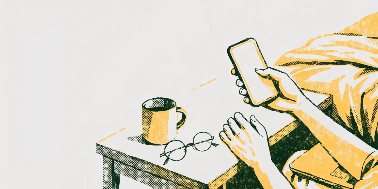 A person's hand holds a smartphone with a blank softly glowing screen over a bedside table, where a mug of tea and a pair of reading glasses sit, while the other hand rests near a closed laptop.